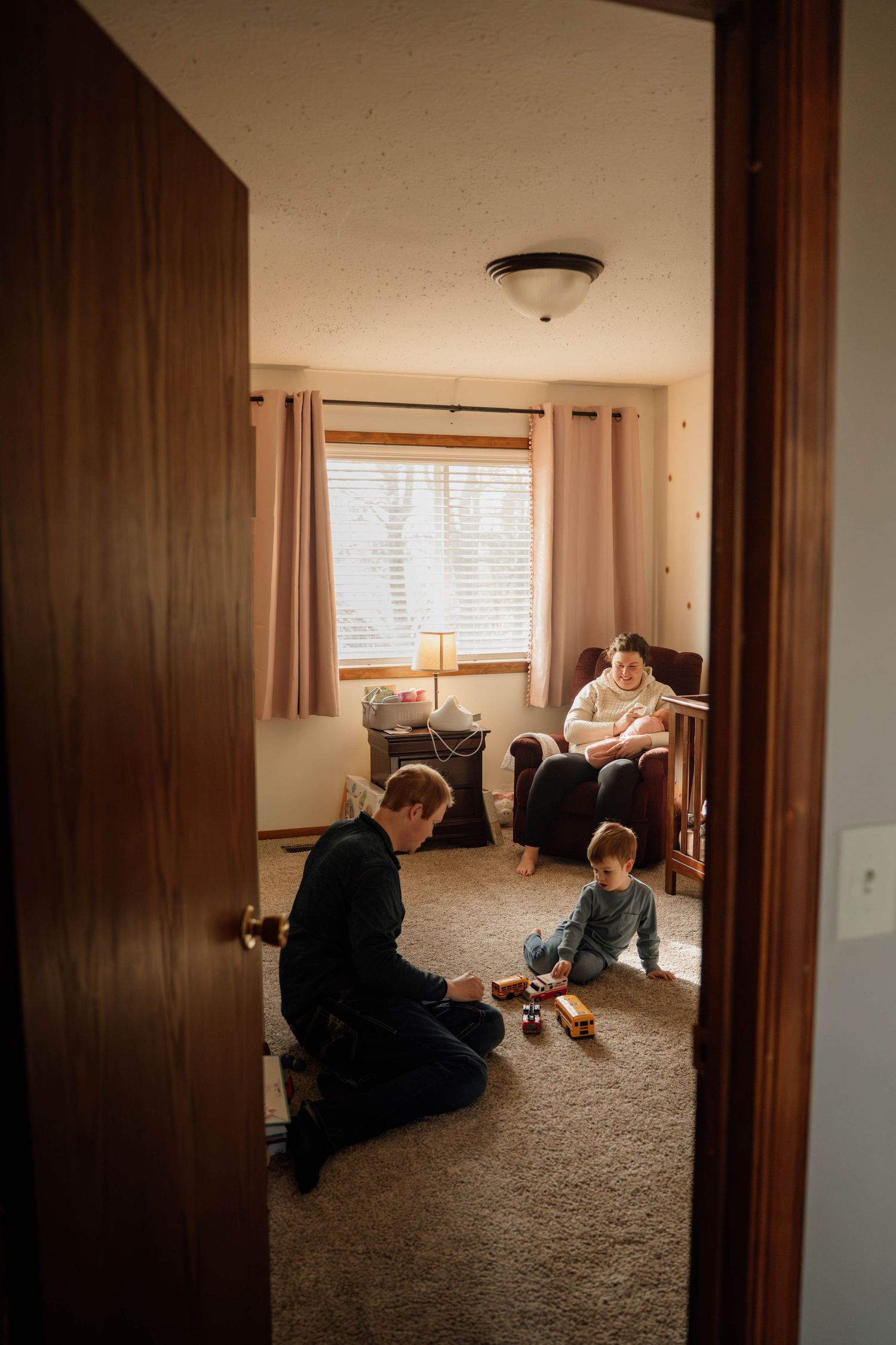Family scene in a cozy nursery with mother holding newborn, child playing on carpet, viewed through doorway.