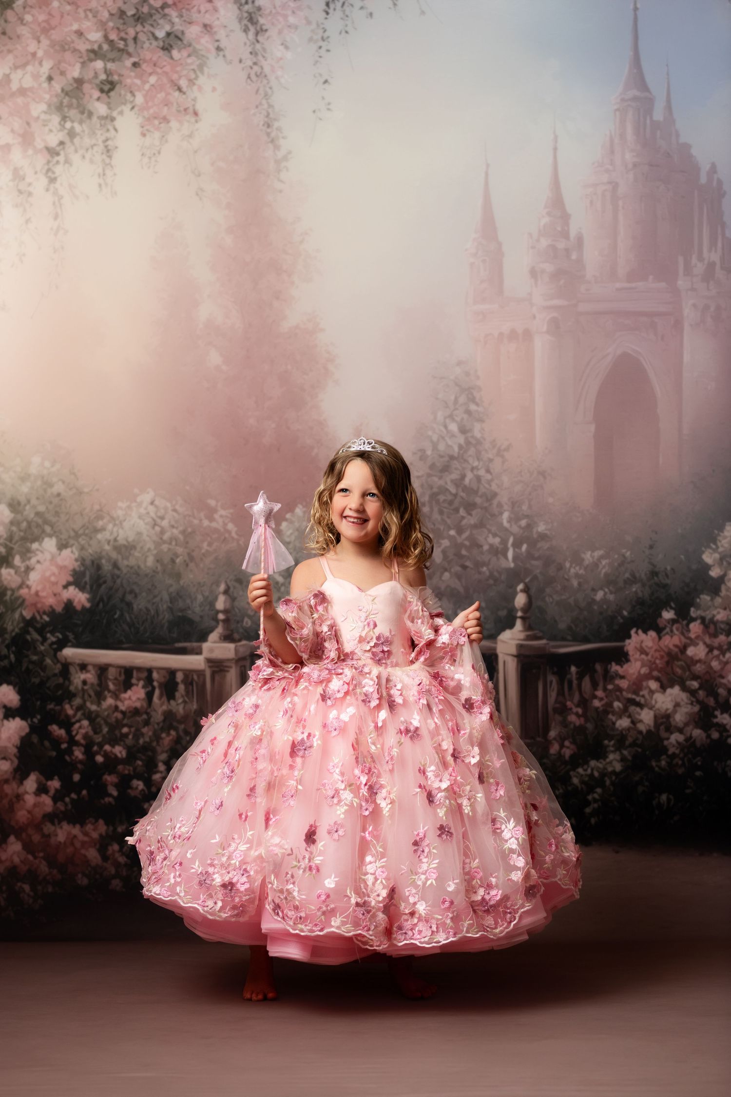Young girl in pink floral princess gown with tiara and wand poses before dreamy castle backdrop.