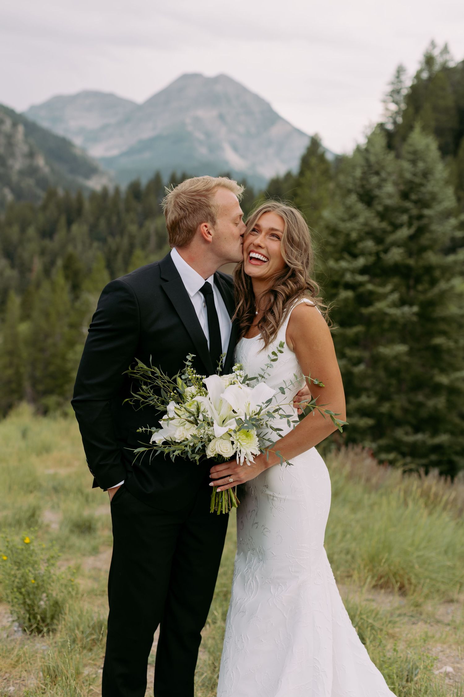 A groom in a black suit kisses his bride in a white gown holding white flowers, with mountain forest backdrop.