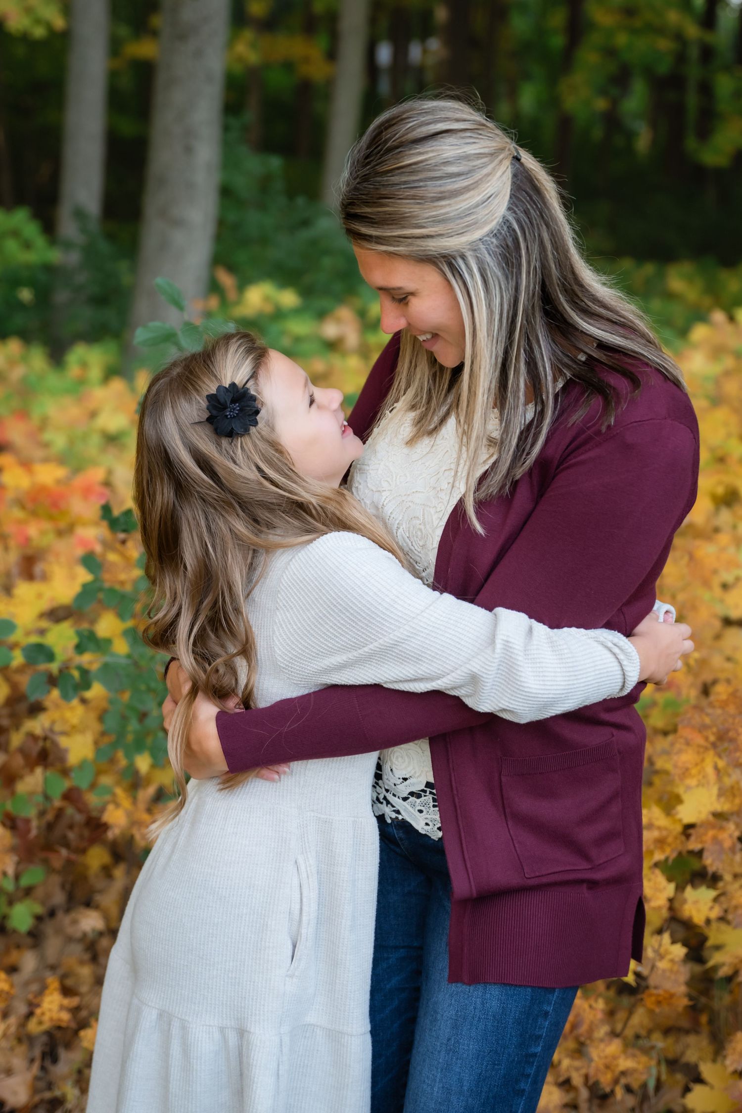 Mother and daughter share a warm embrace outdoors among colorful autumn foliage in a heartfelt fall portrait.