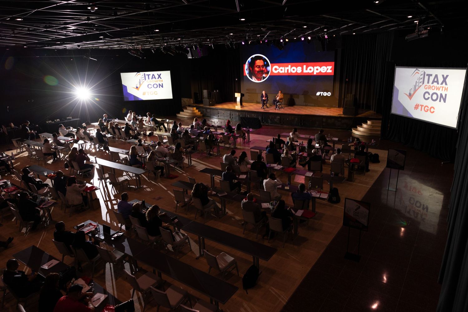 Aerial view of iTax Con conference with attendees seated in a large auditorium watching Carlos Lopez speak on stage.