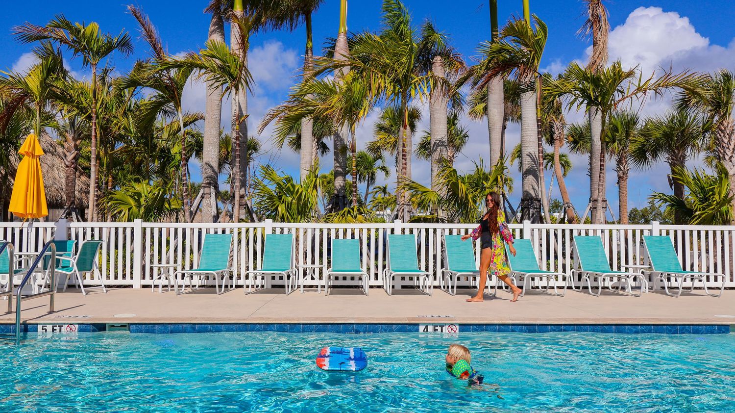Child playing by a tropical resort pool surrounded by palm trees and turquoise lounge chairs on a sunny day.