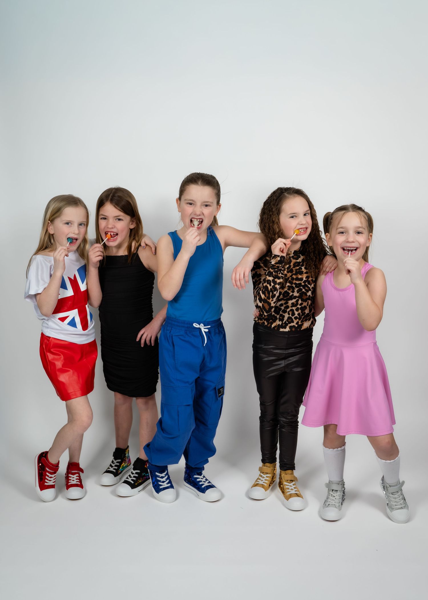 Five children pose playfully in colorful outfits, including a Union Jack skirt, black dress, blue tracksuit, leopard print, and pink dress.
