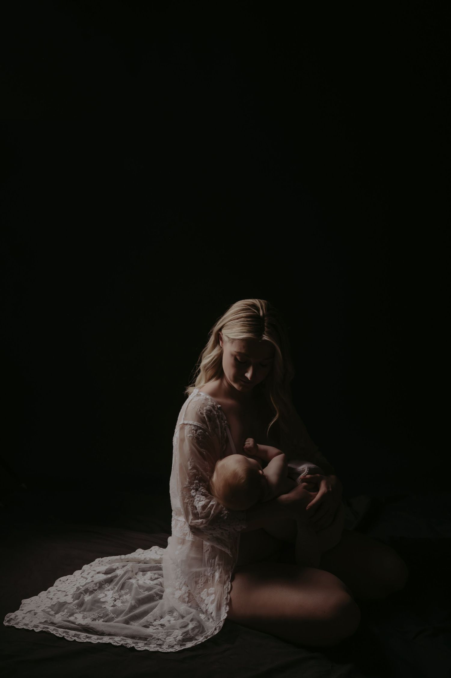 A mother in white lace gown cradles her newborn baby in a dark, moody studio portrait.