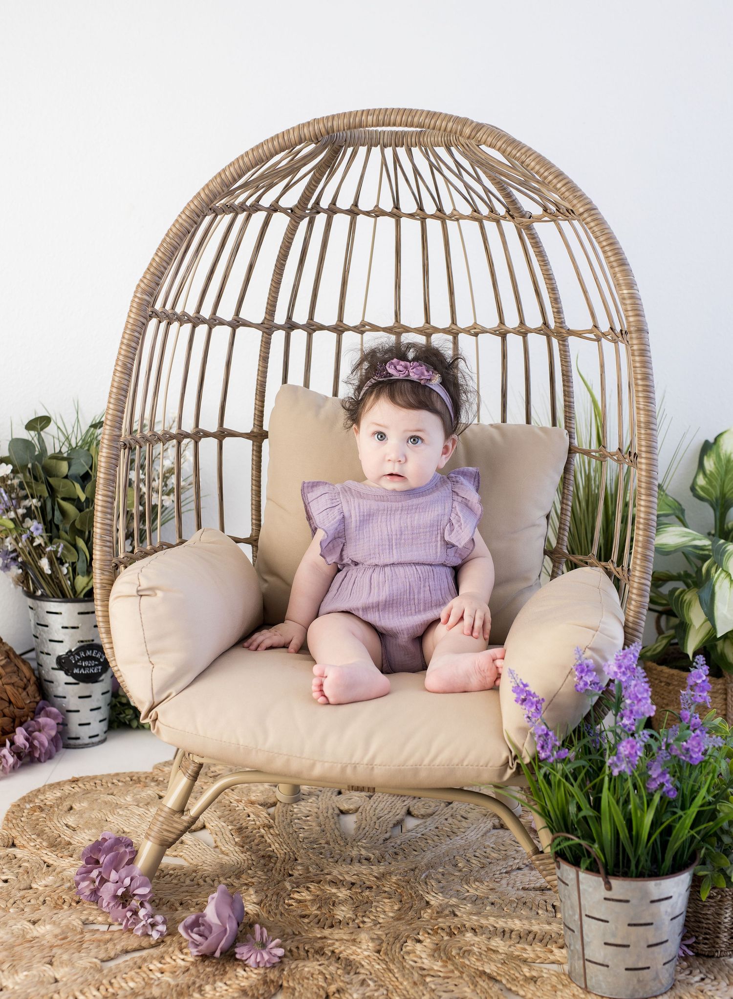 Baby girl in purple romper sits in a wicker egg chair surrounded by lush greenery and purple flowers.