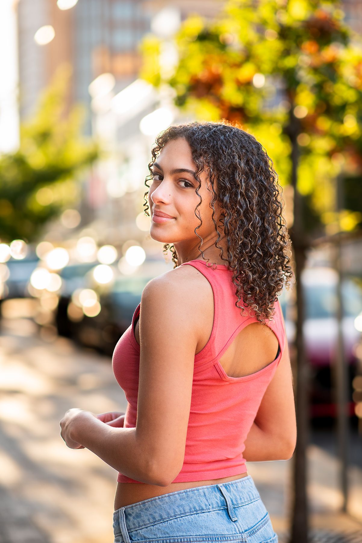 Young woman with curly hair in pink tank top and jeans smiling over shoulder on sunny city street.