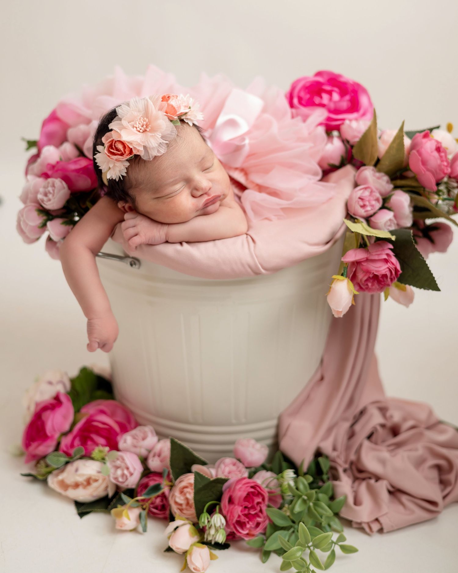 Sleeping newborn baby girl in white bucket surrounded by pink roses wearing floral headband in professional photo.