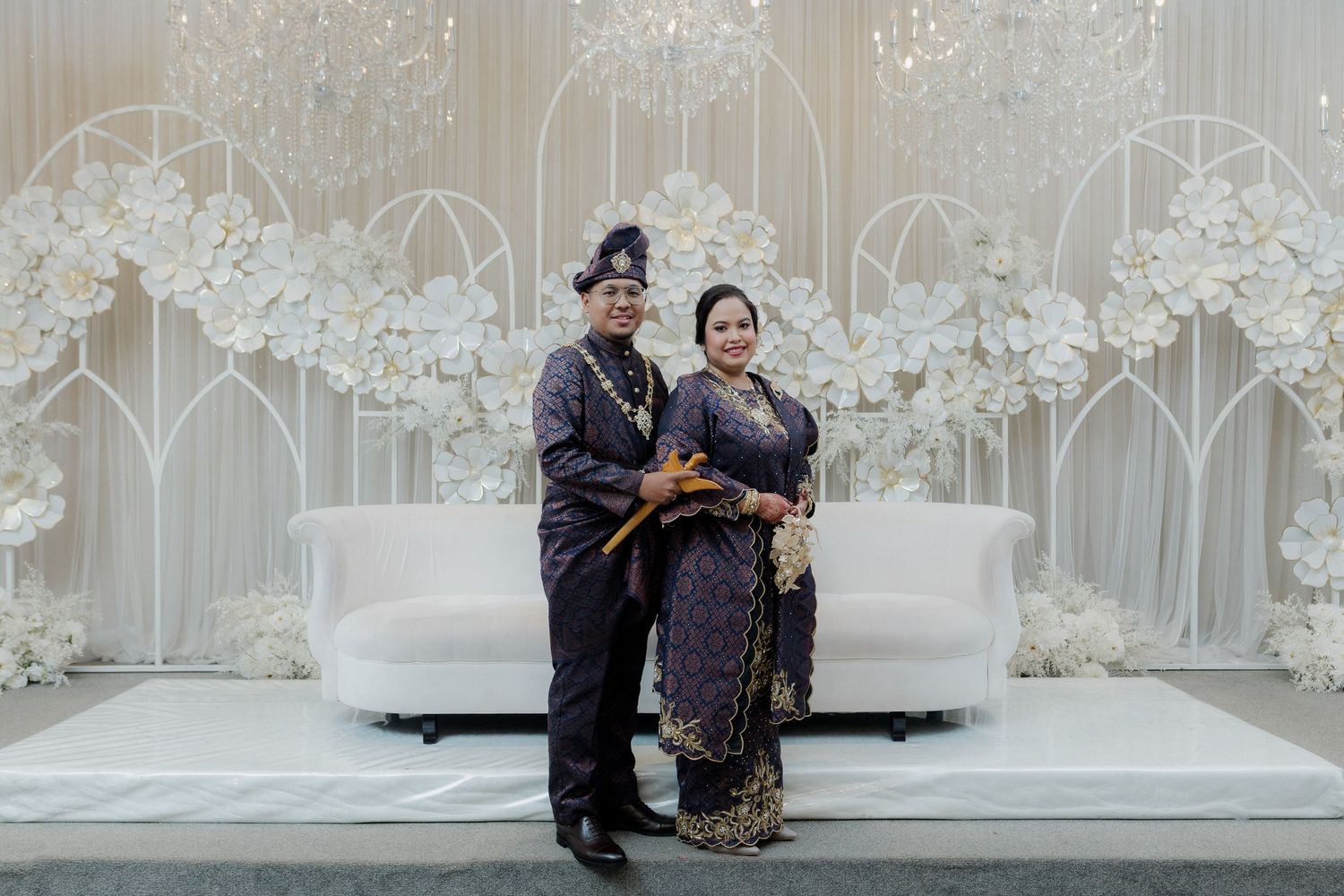 Muslim couple in traditional Javanese wedding attire sitting together at an outdoor ceremony with floral decorations and ocean view.