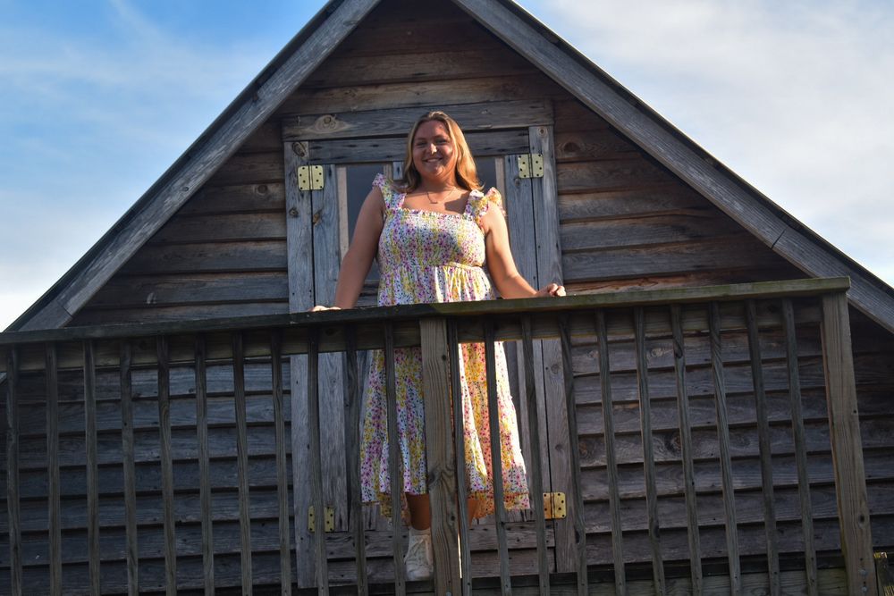 Smiling blonde woman standing on wooden barn balcony in floral sundress.