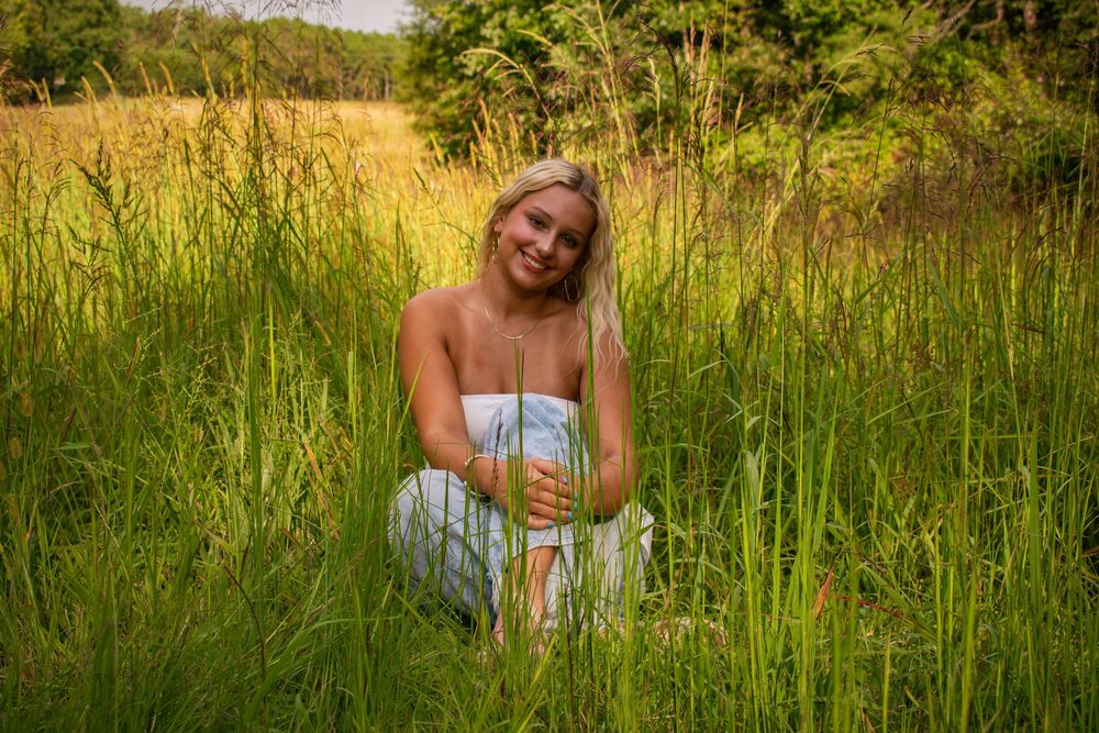 Blonde woman in white dress sitting in tall green grass in a meadow.