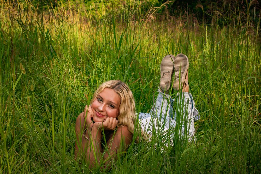 Smiling blonde girl lying in tall green grass with feet up in summer field.