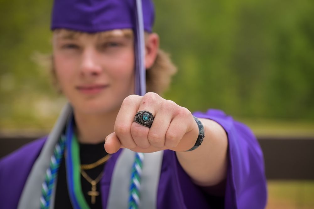 Graduate in purple cap displaying class ring with graduation regalia and cross necklace.