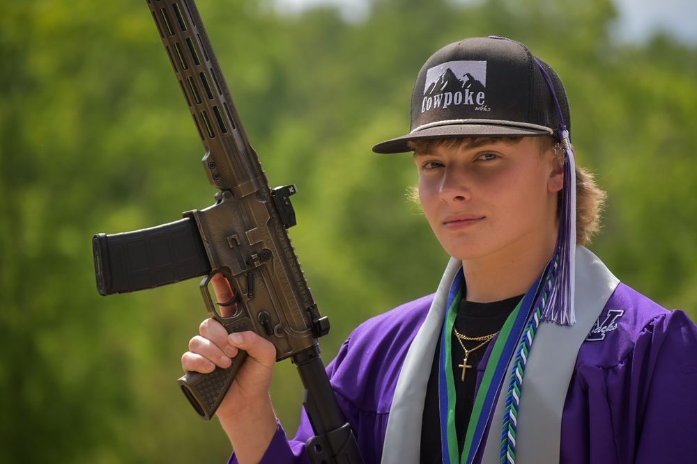 Graduate in purple gown holding rifle wearing black cap and graduation regalia outdoors.