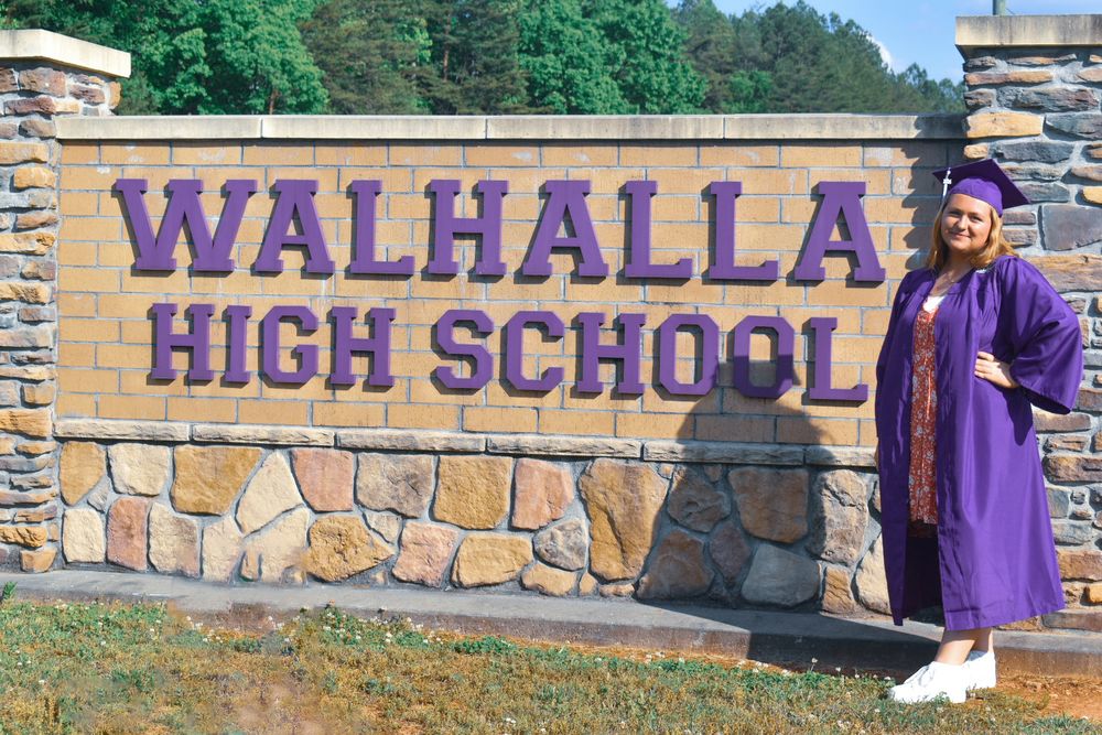 Graduate in purple cap and gown posing beside Walhalla High School sign.