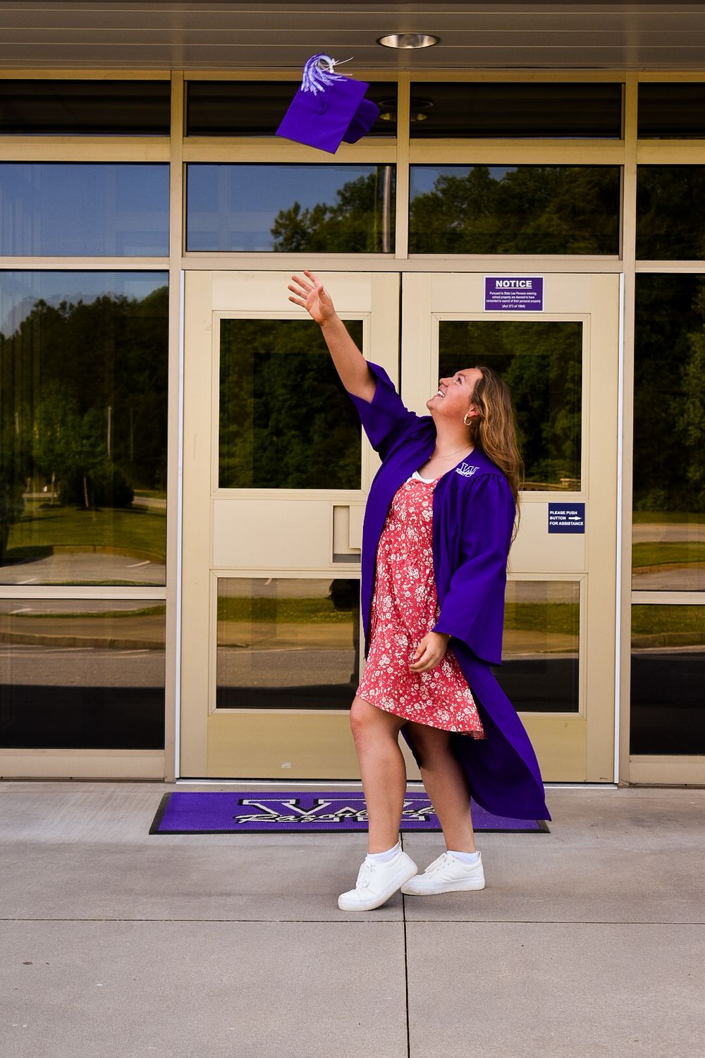 Graduate in purple cap and gown tossing cap in the air outside school entrance.