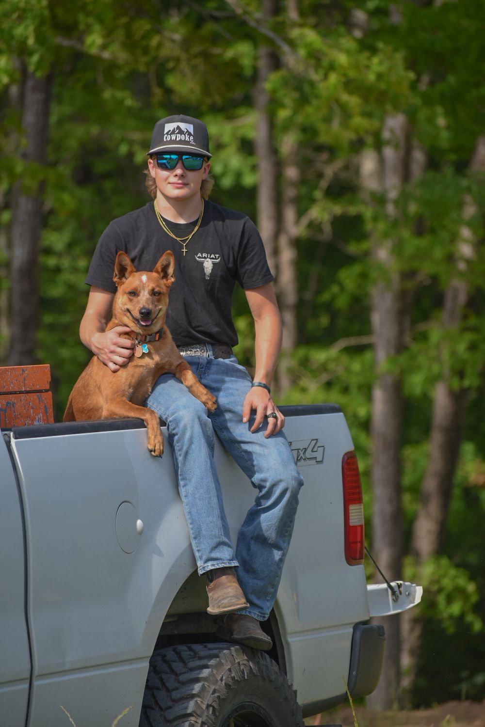 Young man sitting on a truck tailgate with a red dog in a wooded area.