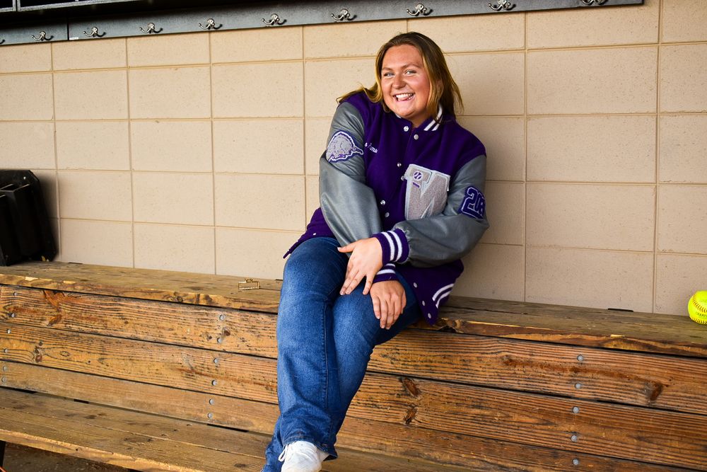 Young woman laughing in a purple varsity jacket sitting on a wooden bench outdoors.