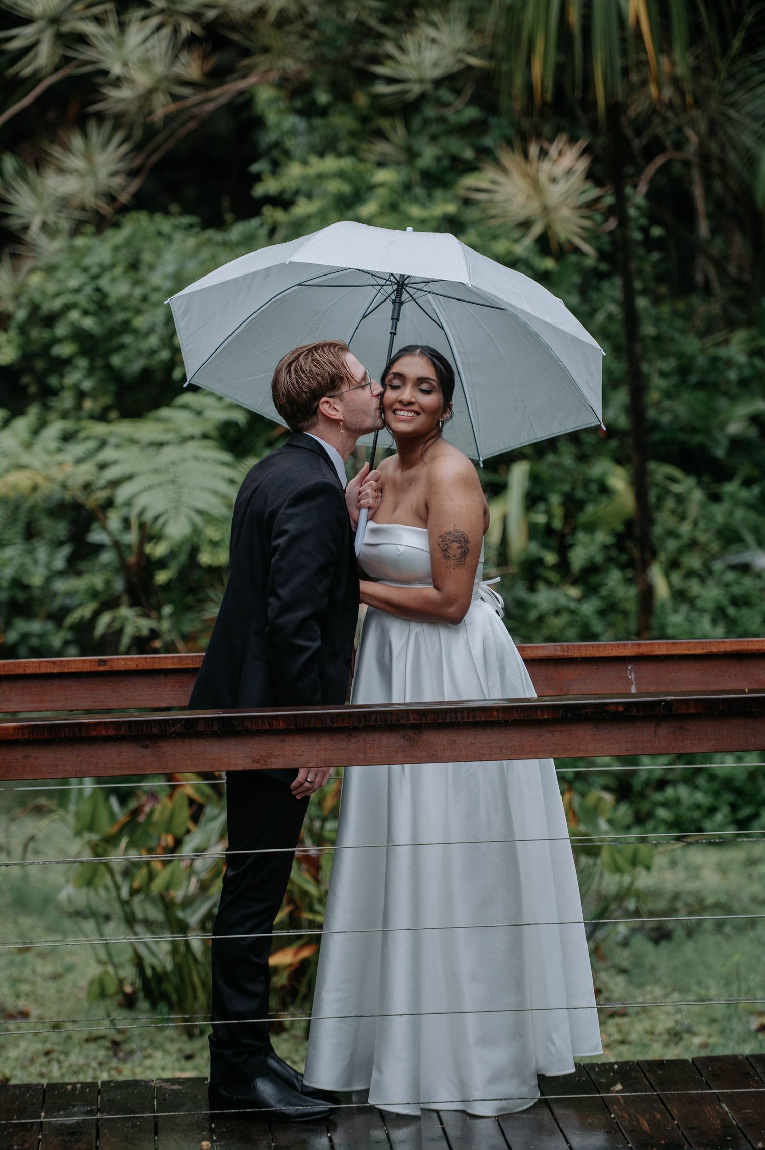 Bride and groom share a kiss under a white umbrella on a rainy wedding day, surrounded by lush greenery.