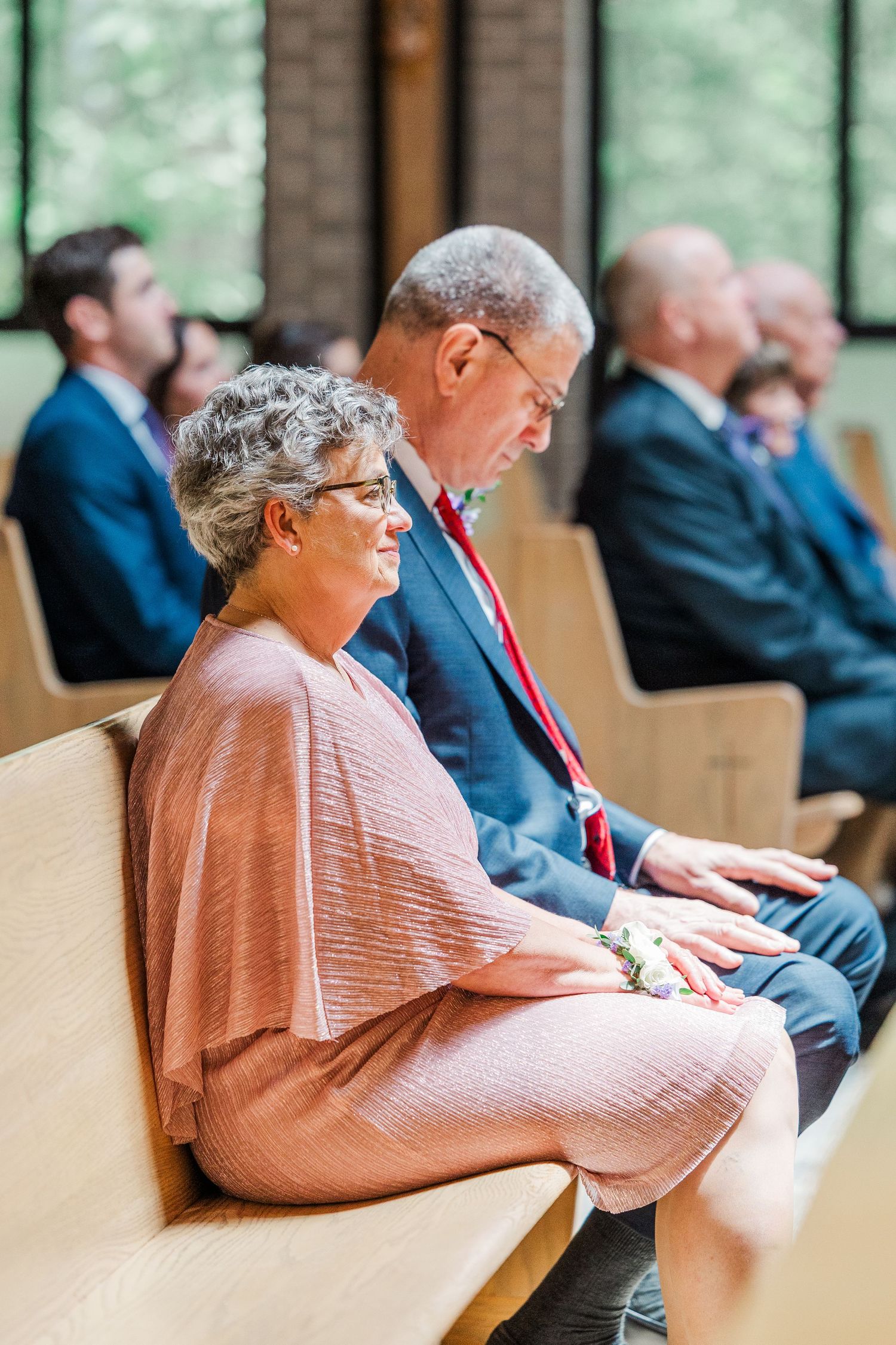 Elderly couple seated in church pew during wedding ceremony, woman in pink dress with corsage, man in blue suit.