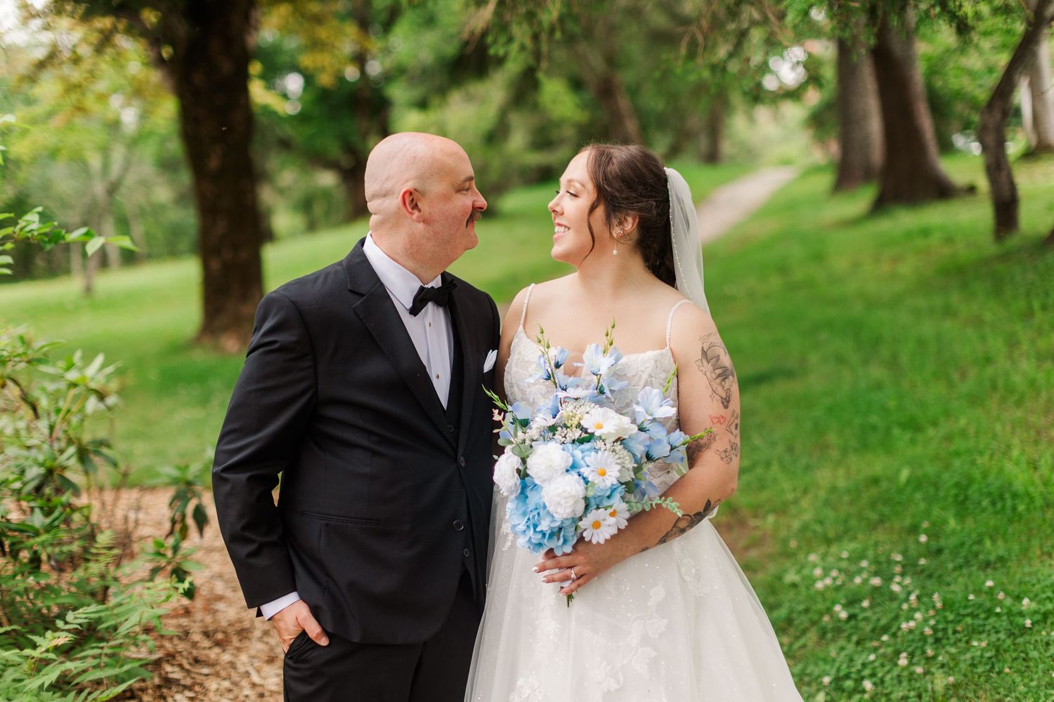 Bride and groom share a romantic moment outdoors, she holds a blue and white bouquet, he wears a black tuxedo.
