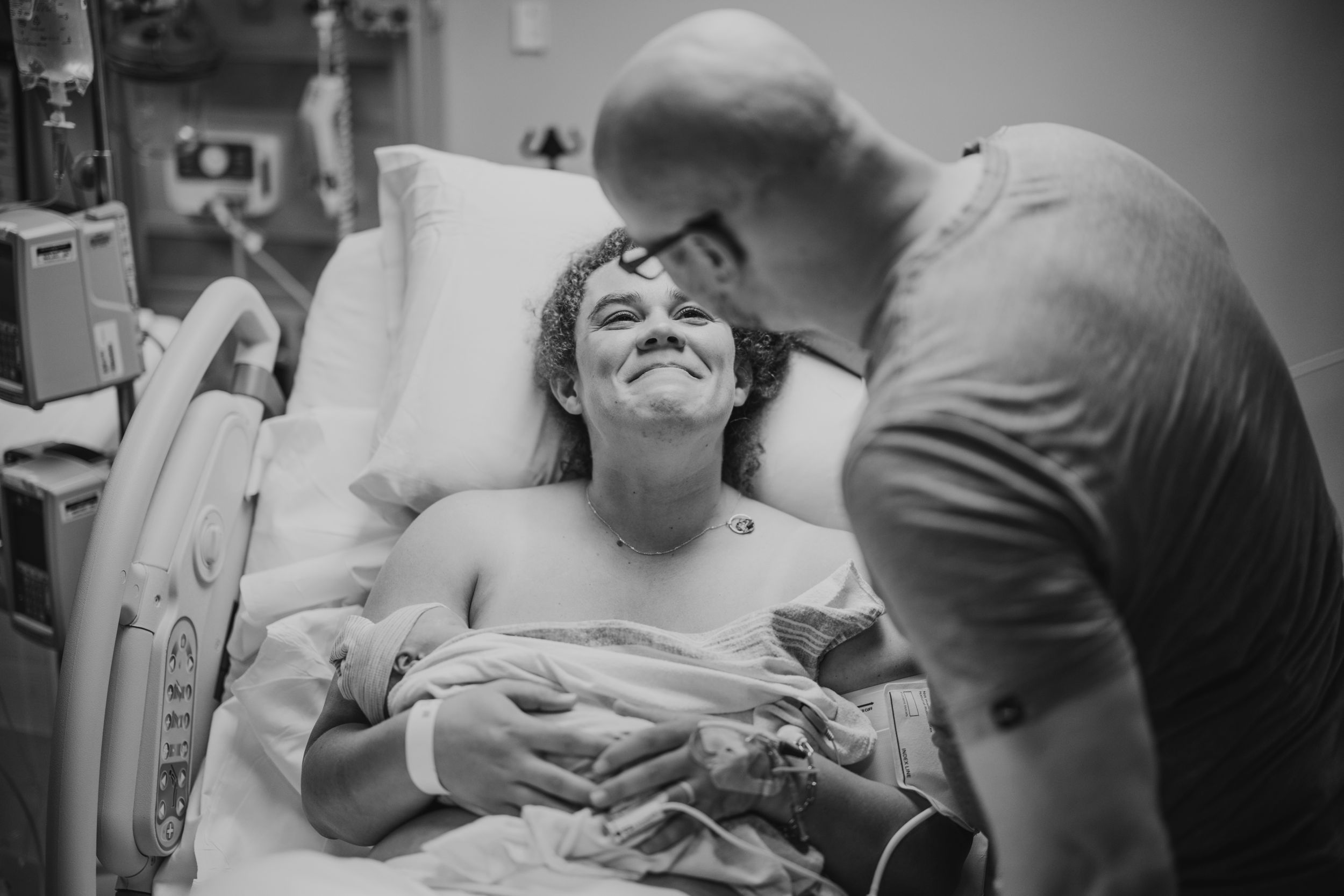 Black and white photo of a new mother smiling in hospital bed holding newborn while partner leans in tenderly.