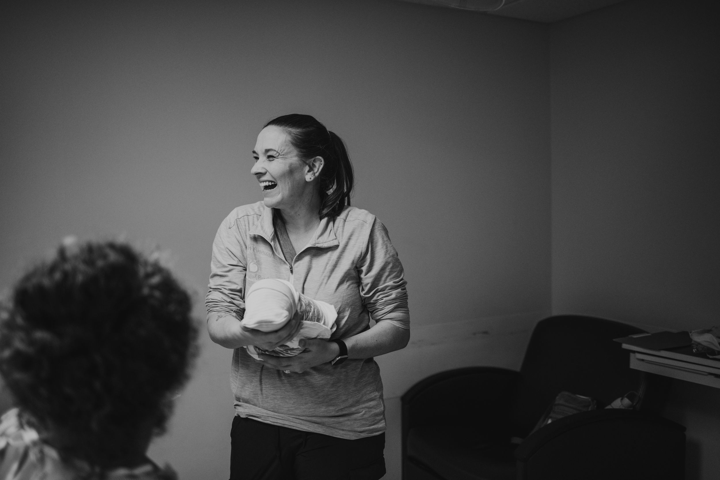 Black and white photo of a smiling woman holding items while presenting in an office setting.