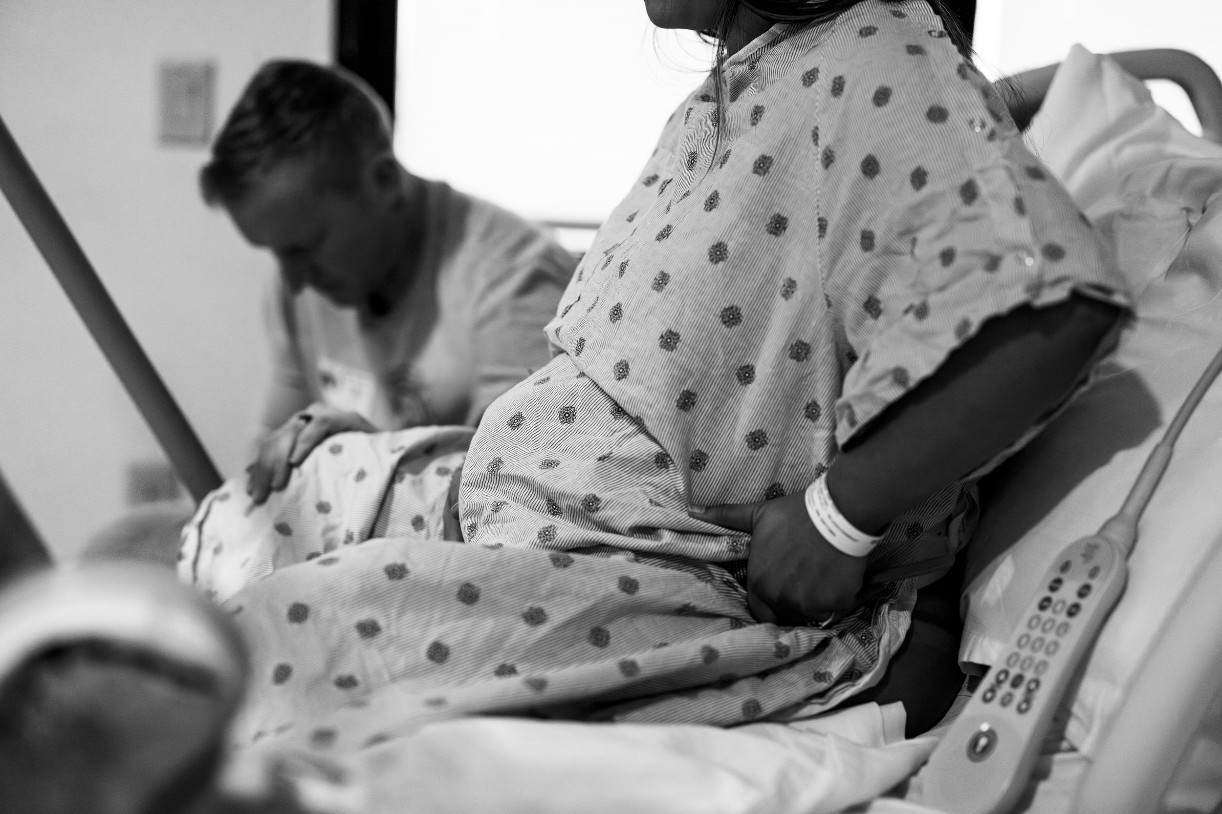 Black and white photo of a pregnant woman in hospital gown sitting in a medical bed during labor.