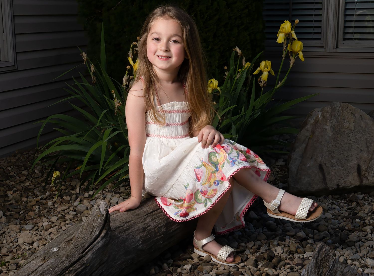 Young girl in white floral dress sitting on a log outdoors, smiling, with yellow iris flowers in background.