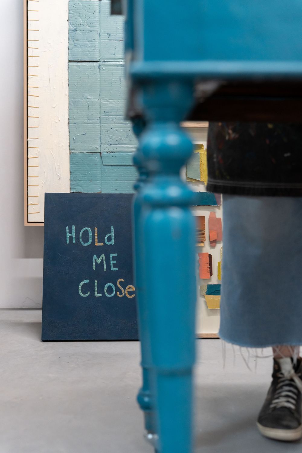 Blue art studio with a dark sign reading Hold Me Close leaning against a teal painted brick wall.