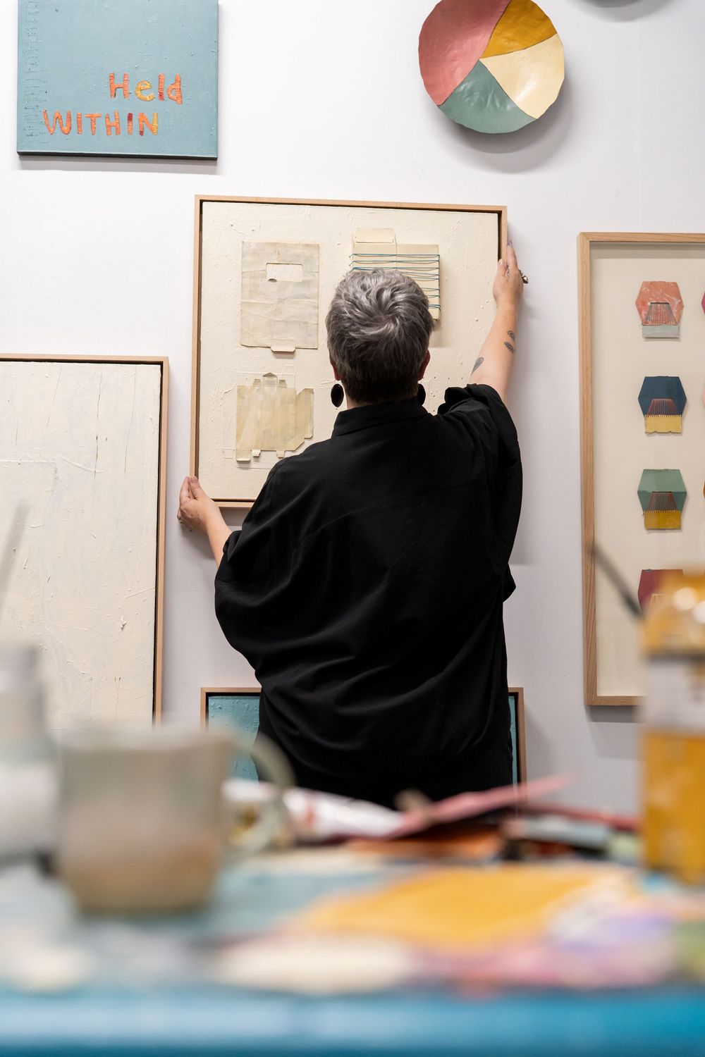 Artist arranging framed artwork on a white gallery wall surrounded by colorful paintings and art supplies.