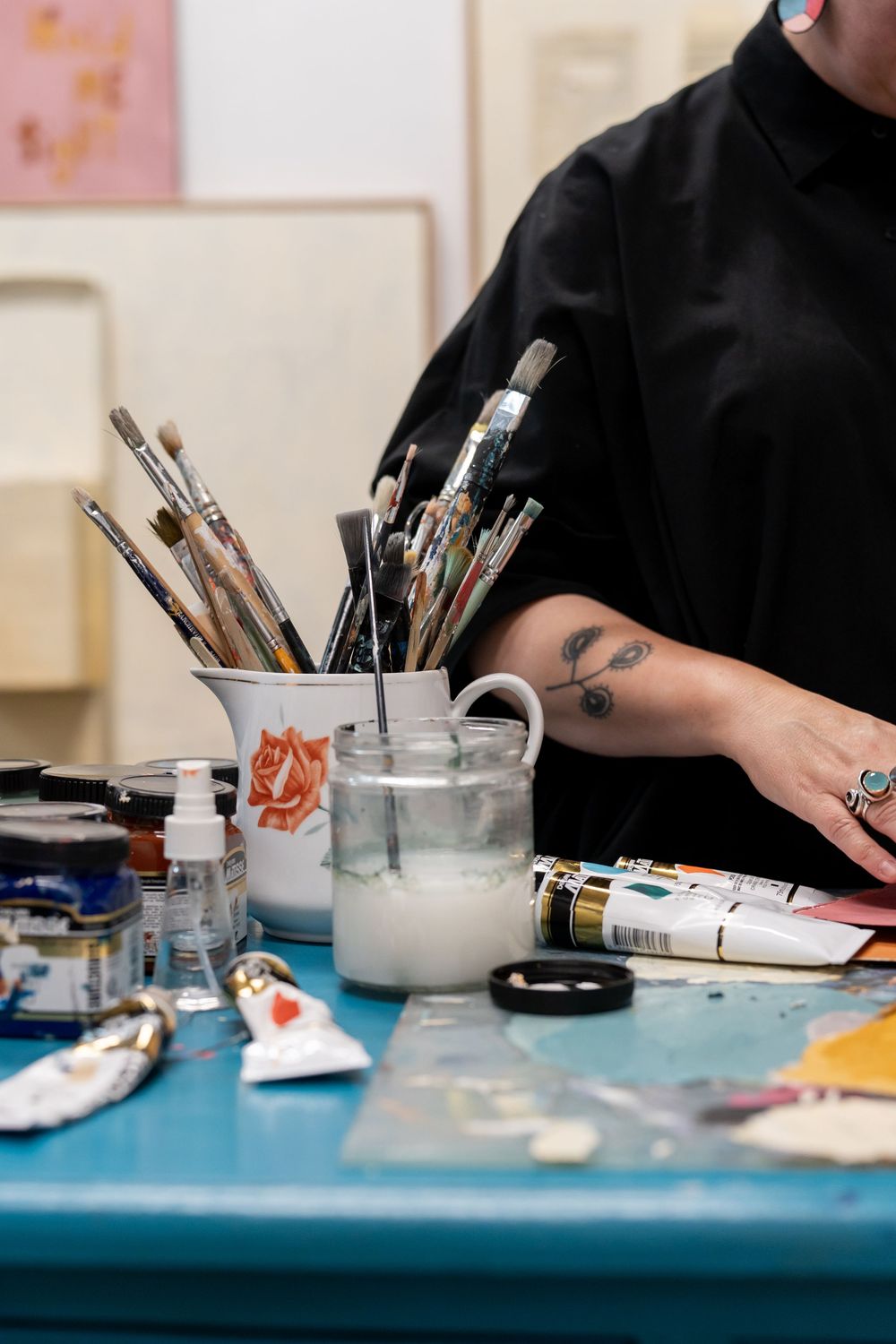 Artist with floral tattoo reaches over a blue table with paint tubes, brushes in a ceramic jug, and art supplies in a studio.