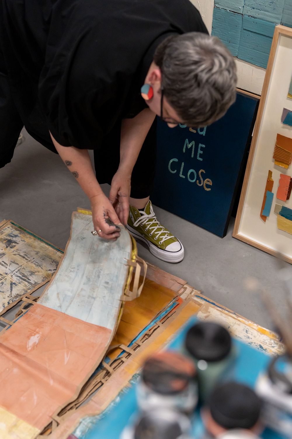 Artist in black outfit and olive Converse sneakers working on a painting on the studio floor.