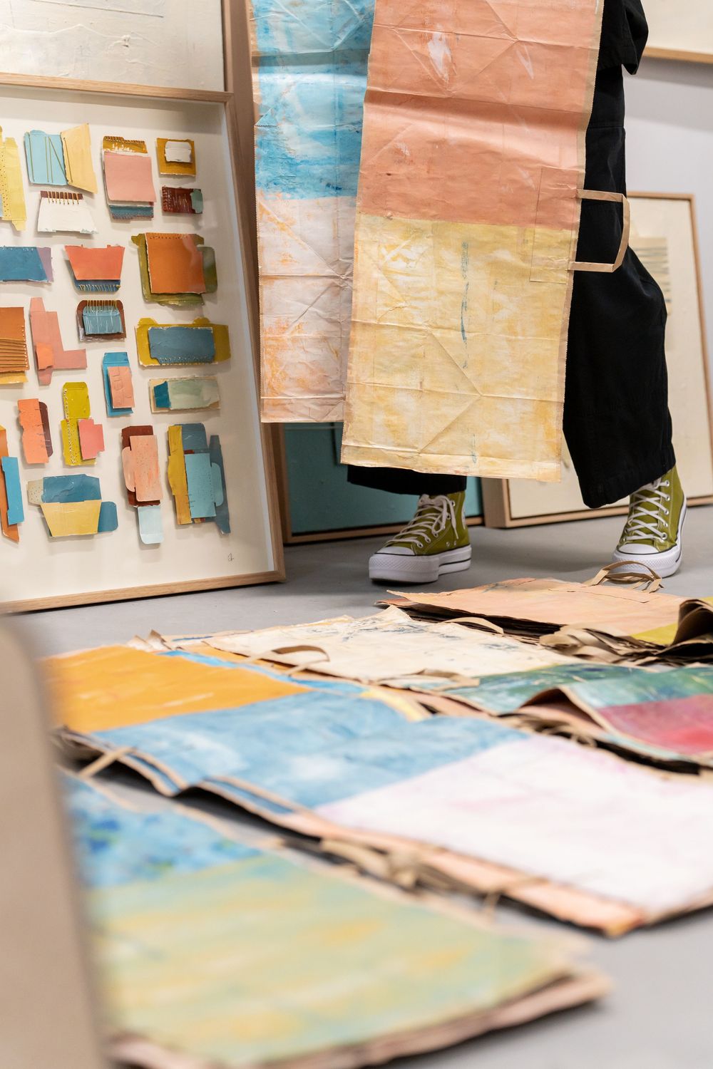 Artist standing among colorful abstract paintings and mixed media artworks displayed on floor and walls in a studio.