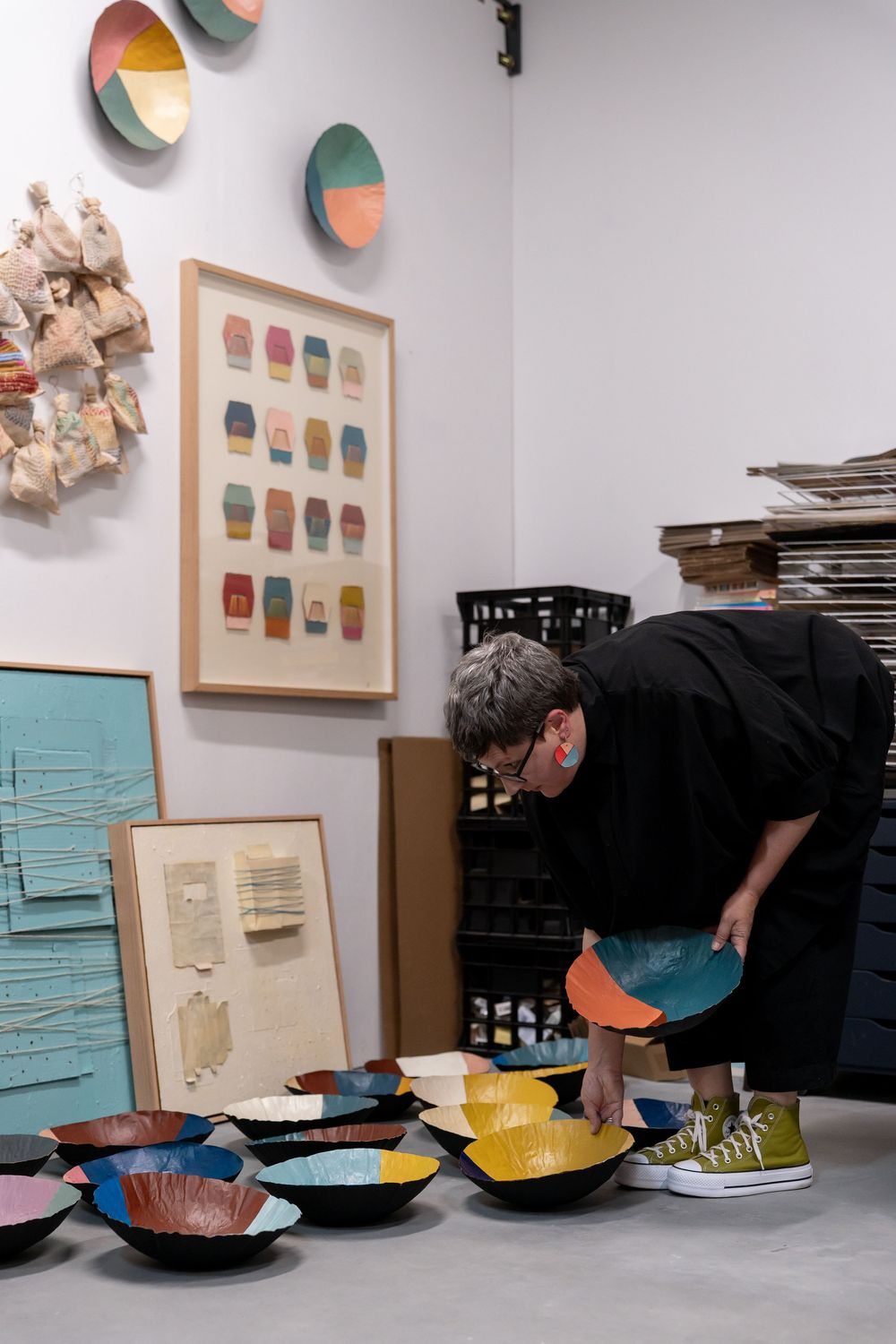 Artist arranging colorful handmade ceramic bowls on a table in an art studio with framed artwork on the wall.