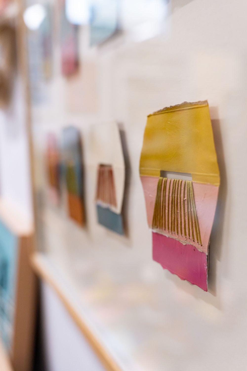 Colorful abstract art pieces mounted on a white gallery wall, featuring pink, yellow, and gold textured mixed media works.