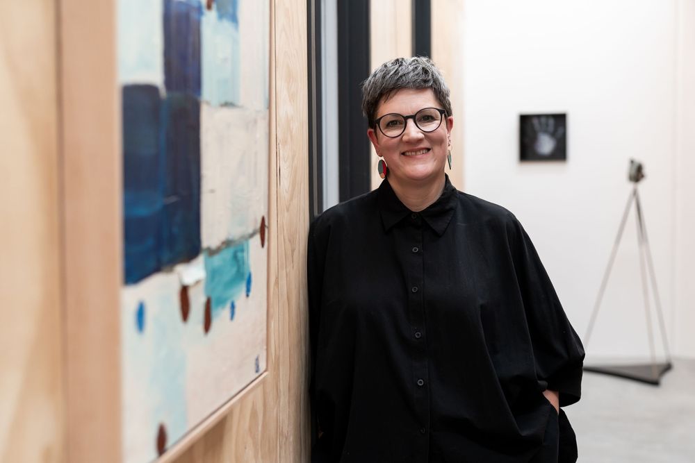 Woman in black outfit smiling in art gallery surrounded by contemporary paintings and sculptures.