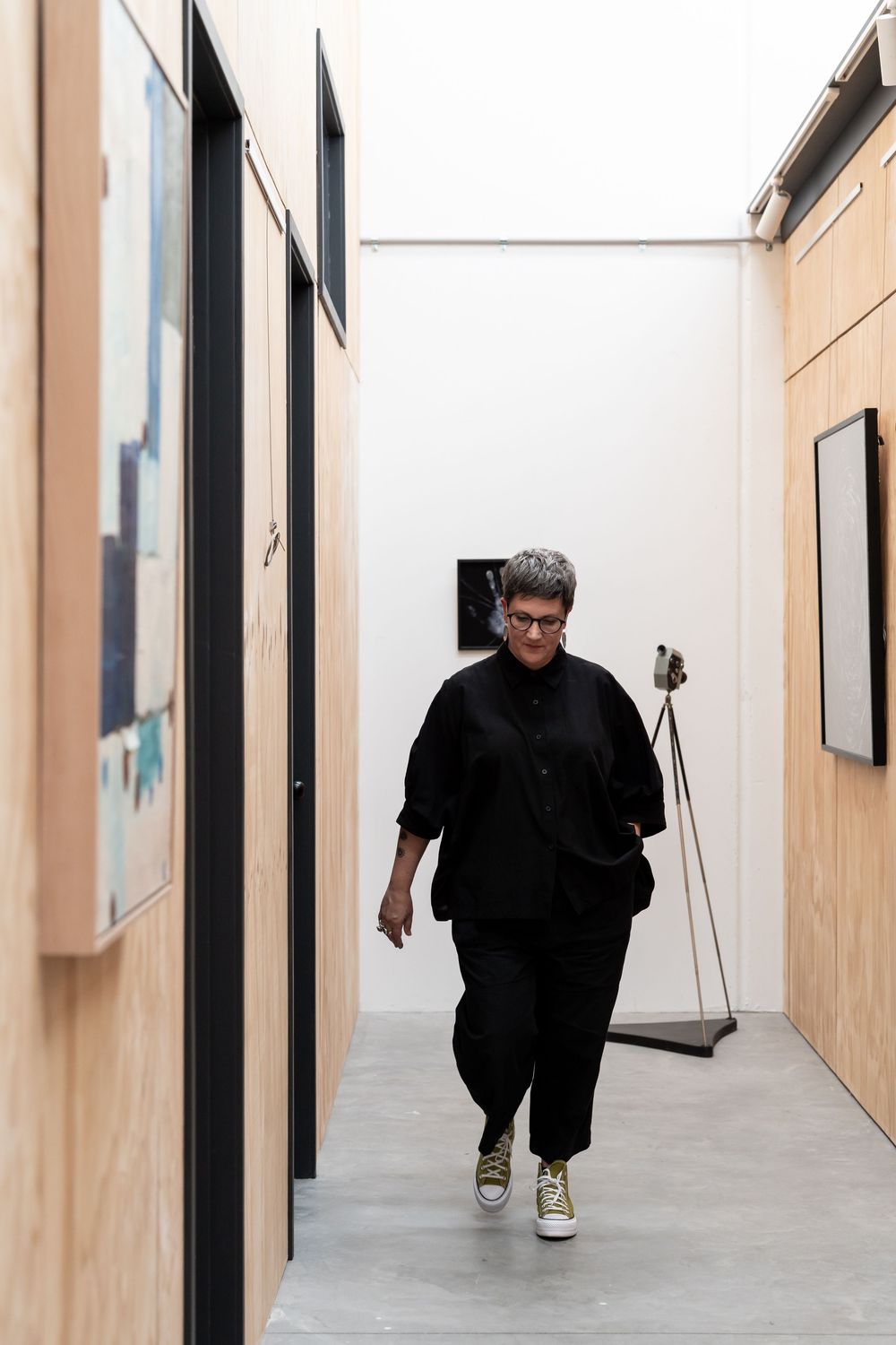 Person in black outfit walking through a modern art gallery corridor with paintings on wooden walls.