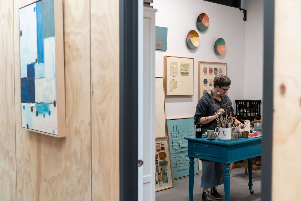 Artist working at a teal desk in a creative studio with abstract paintings and decorative wall art.