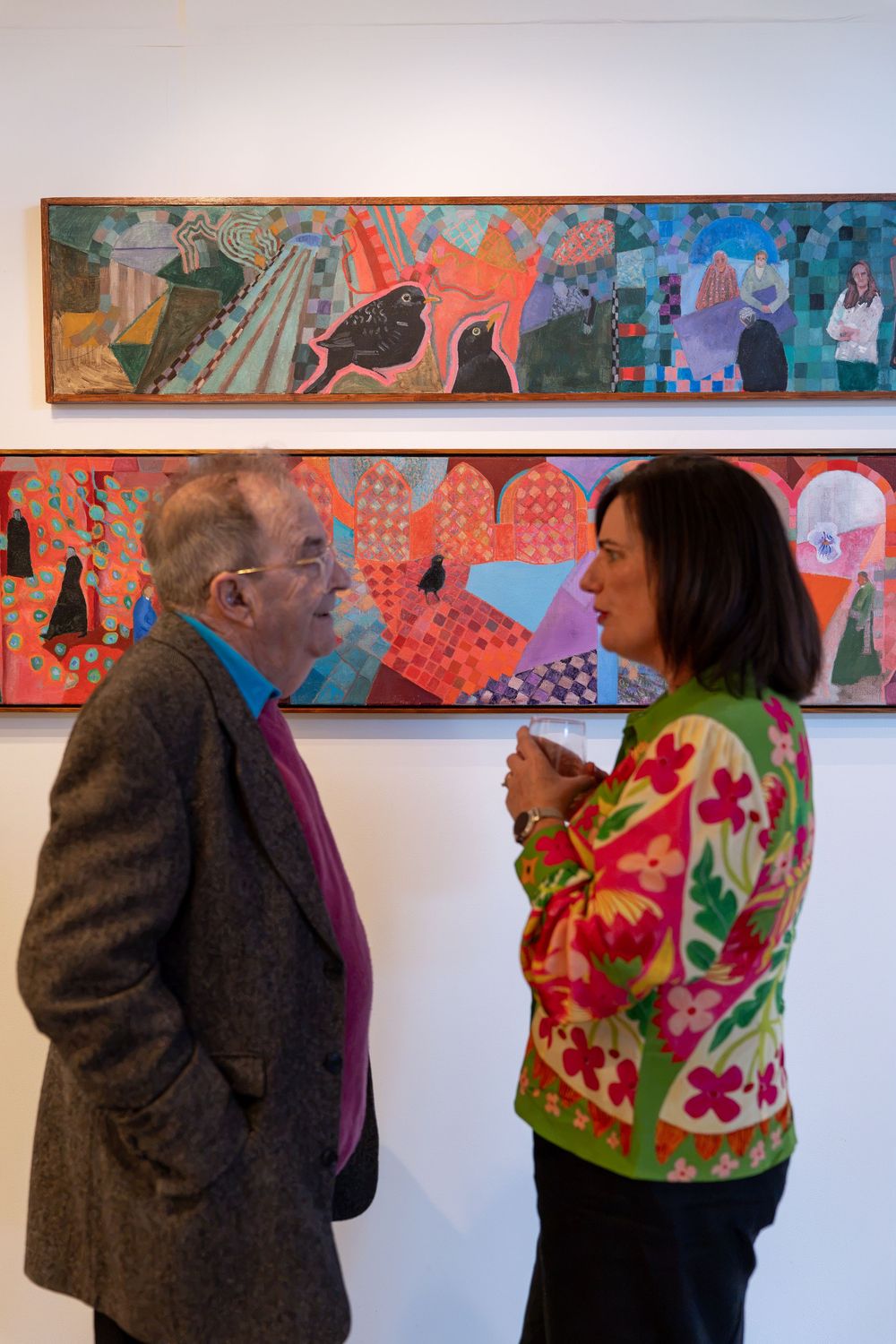 Two people conversing in an art gallery in front of colorful abstract paintings on a white wall.