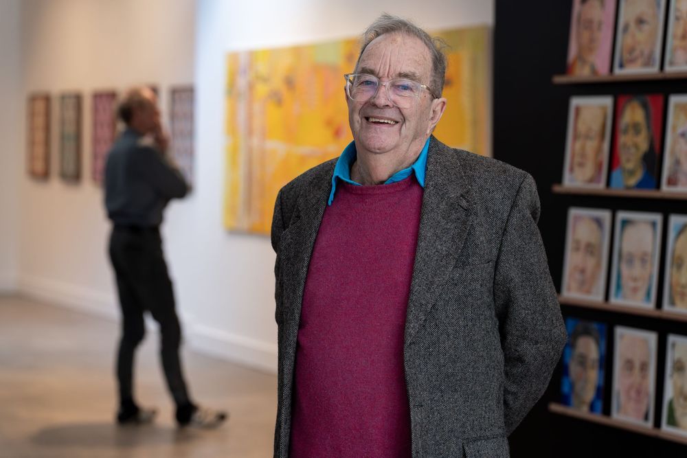 Smiling elderly man in red sweater and gray blazer stands in art gallery with colorful portrait paintings on display.