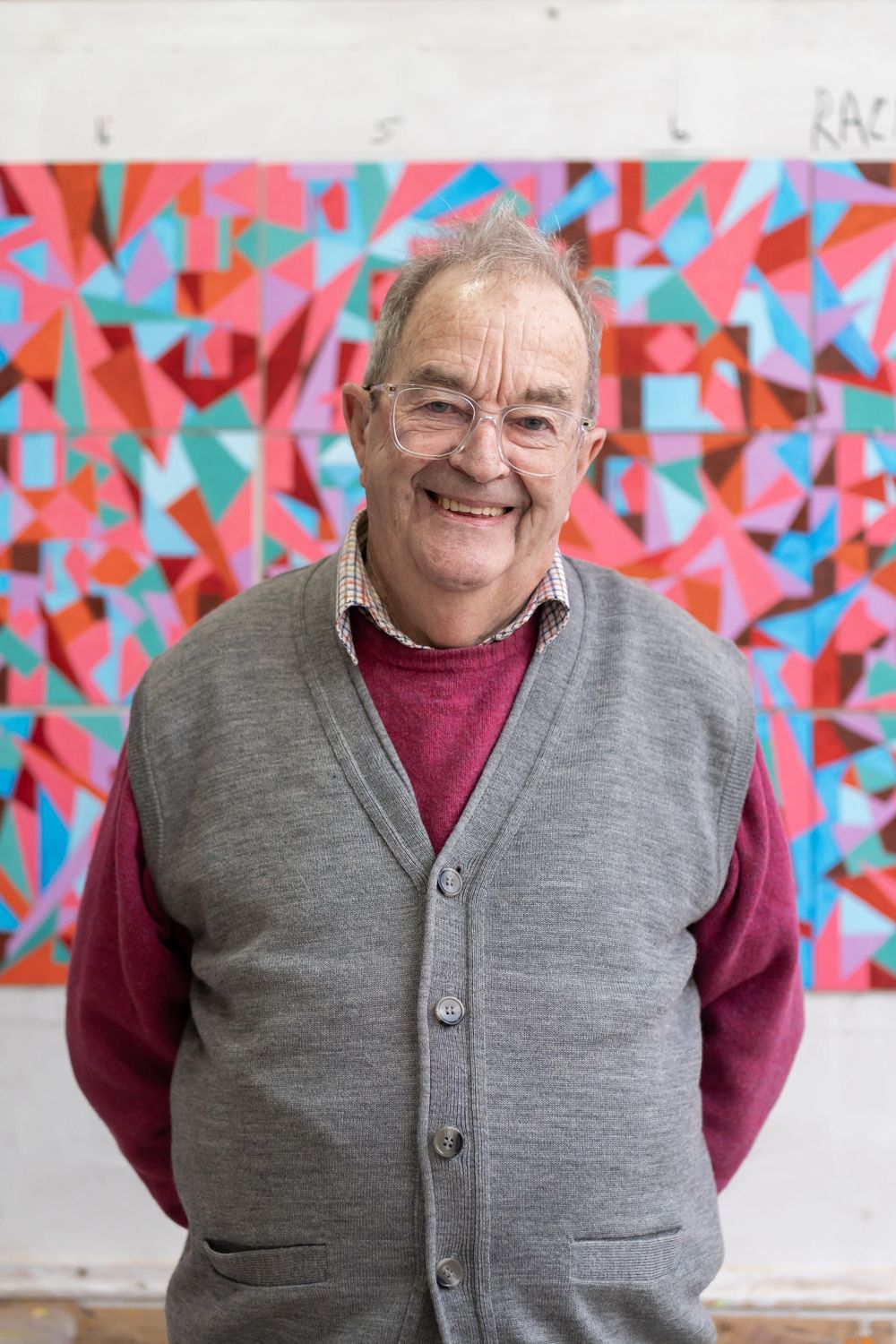 Smiling elderly man in gray vest and red sweater standing before a colorful geometric abstract painting.