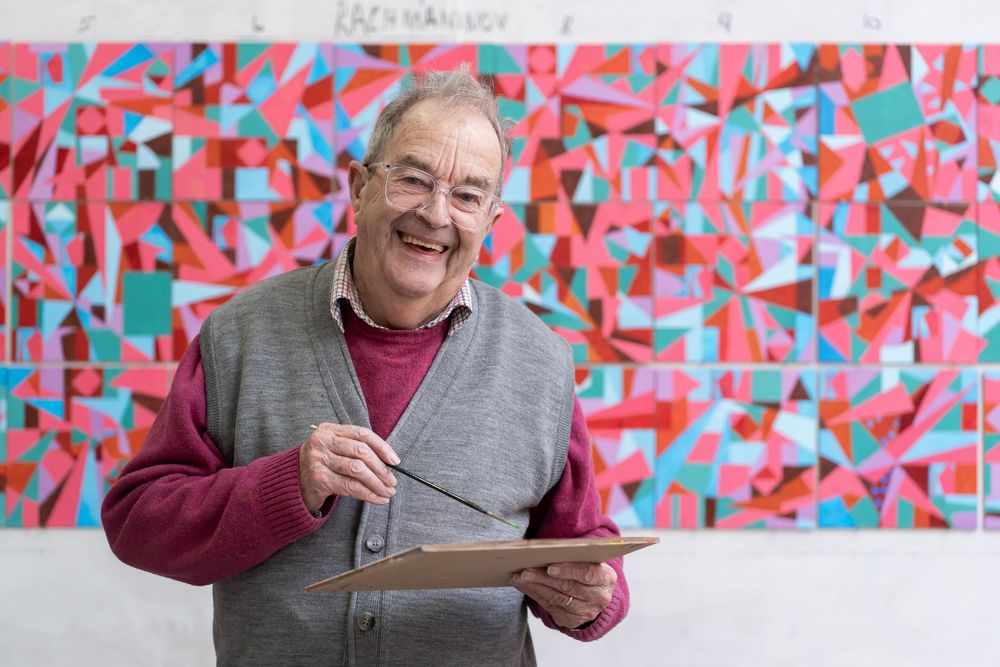 Smiling elderly male artist holding a paintbrush and palette, standing in front of a vibrant colorful abstract painting.