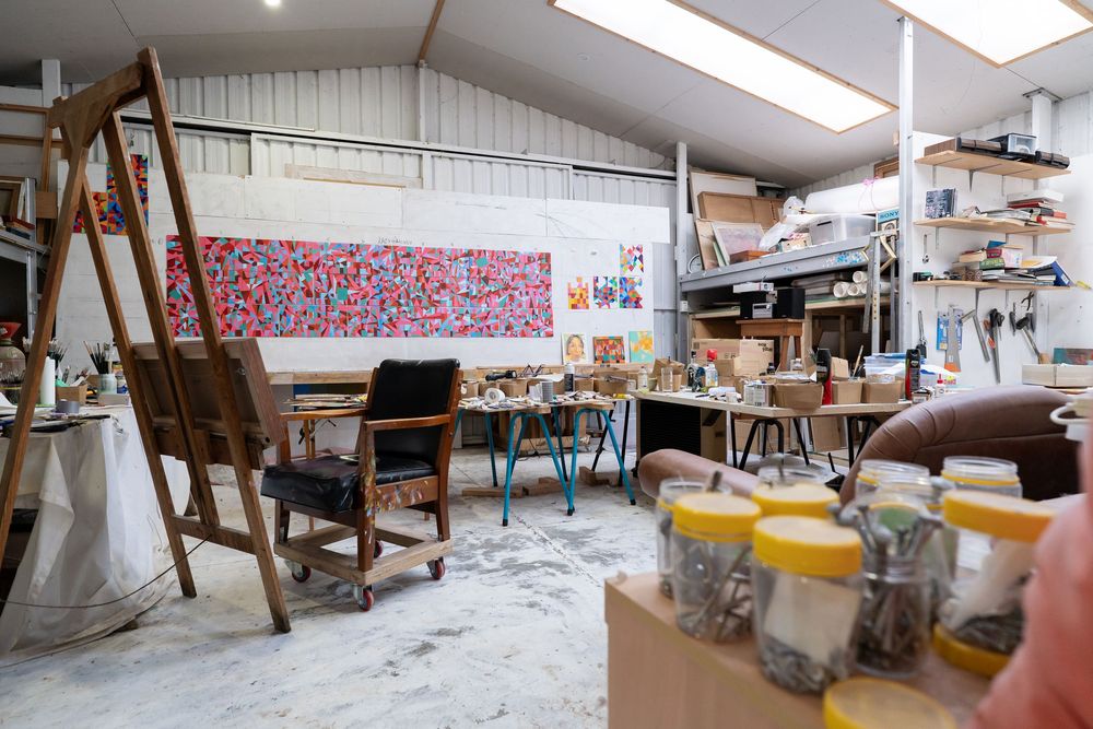 A cluttered artist studio with an easel, colorful paintings, wooden chairs, and art supplies on a paint-splattered floor.