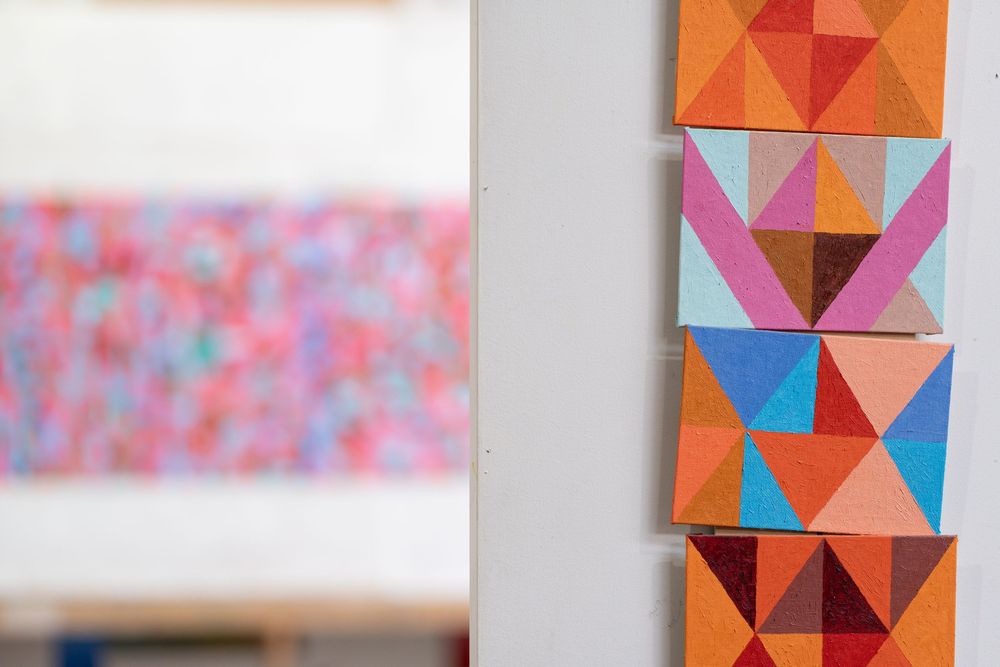 Colorful geometric triangle pattern paintings in orange, pink, and blue displayed on a white wall in an art studio.