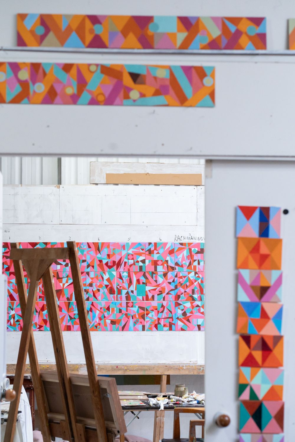 Colorful geometric abstract paintings displayed in an artist studio with wooden easels and vibrant pink, orange, and blue patterns.
