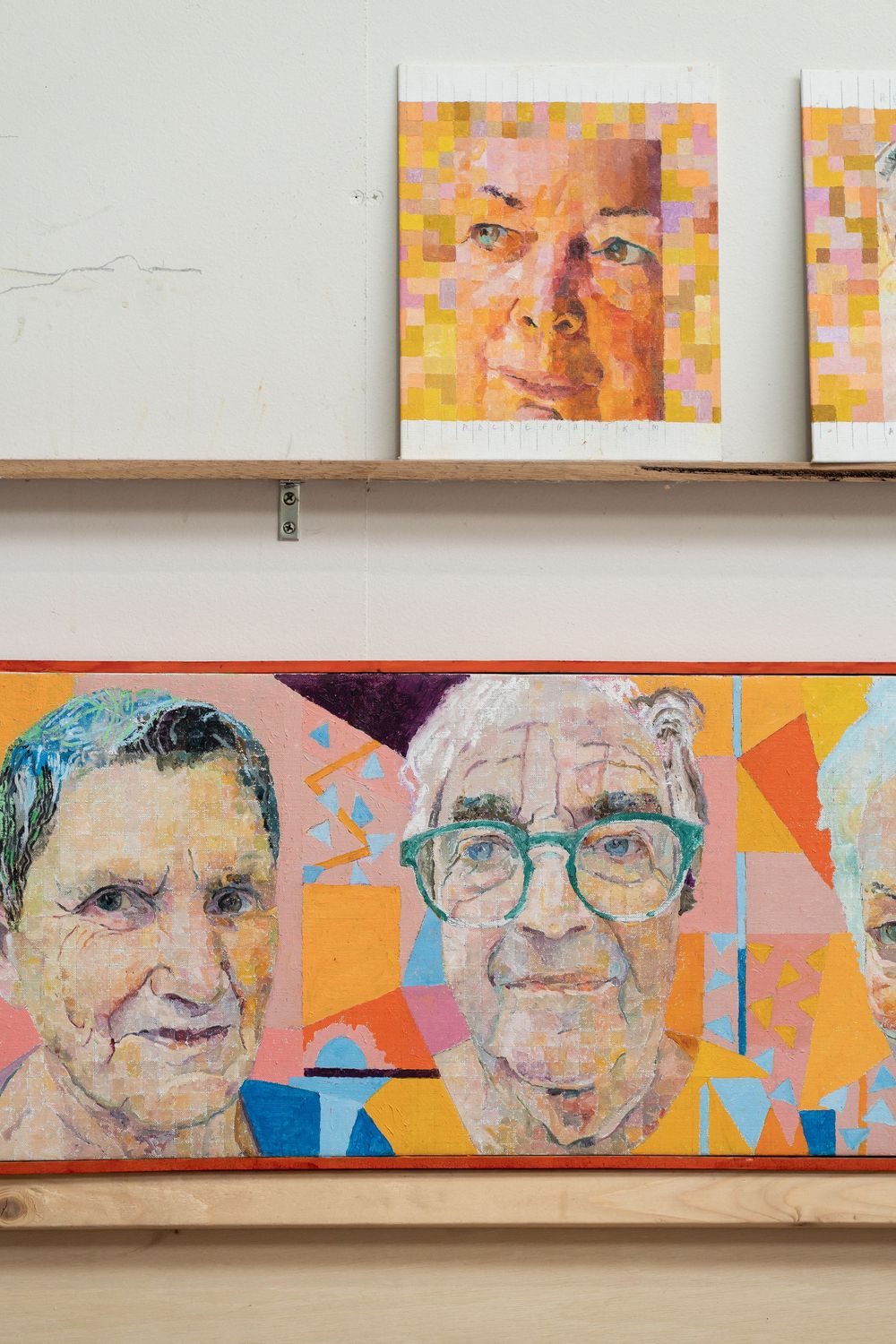 Colorful portrait paintings of elderly people displayed on wooden shelves in an art studio setting.