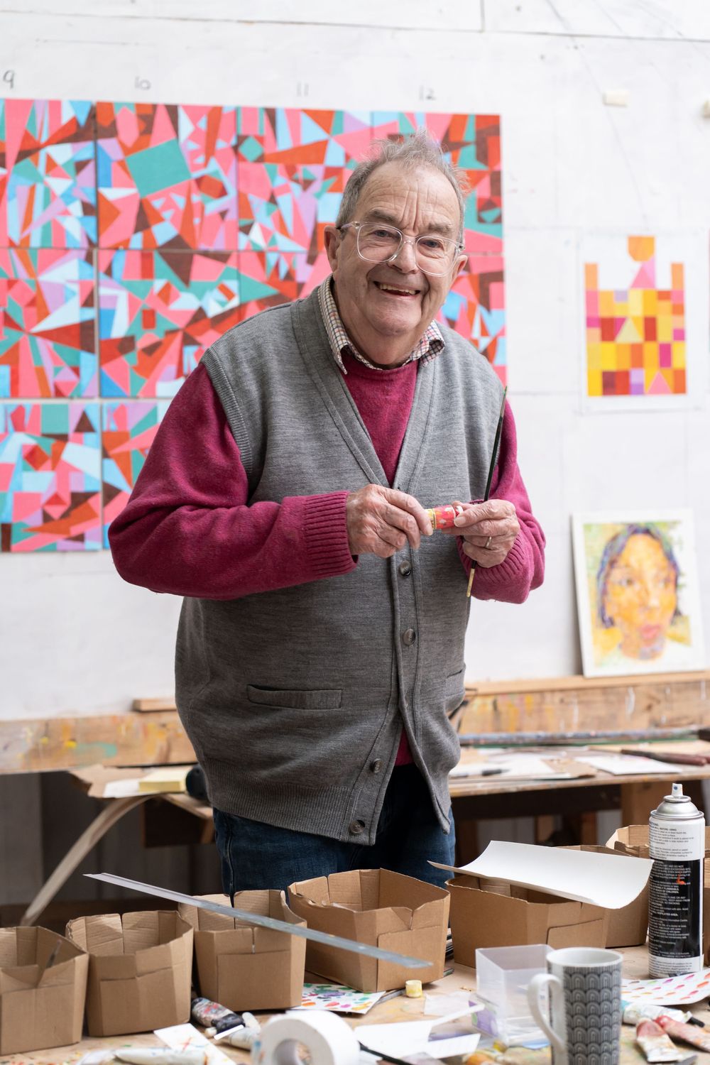 Smiling older man in gray vest stands in art studio with colorful geometric paintings on wall behind him.