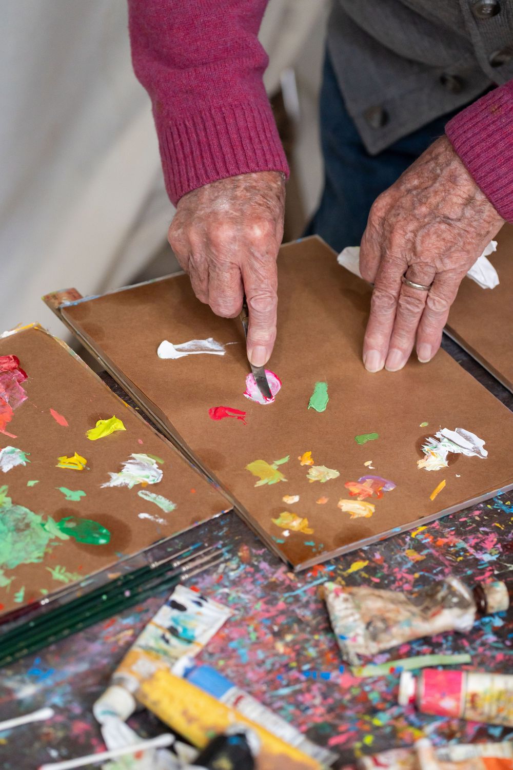 Elderly artist's hands applying colorful paint strokes onto brown canvas board in a creative studio setting.