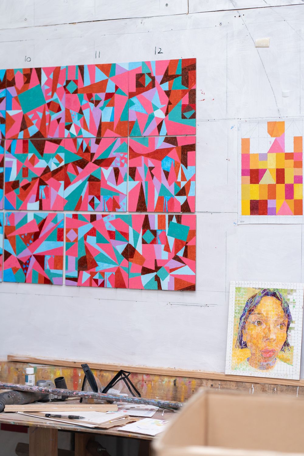 Artist studio wall displaying vibrant geometric abstract painting in pink and teal beside a warm-toned portrait artwork.