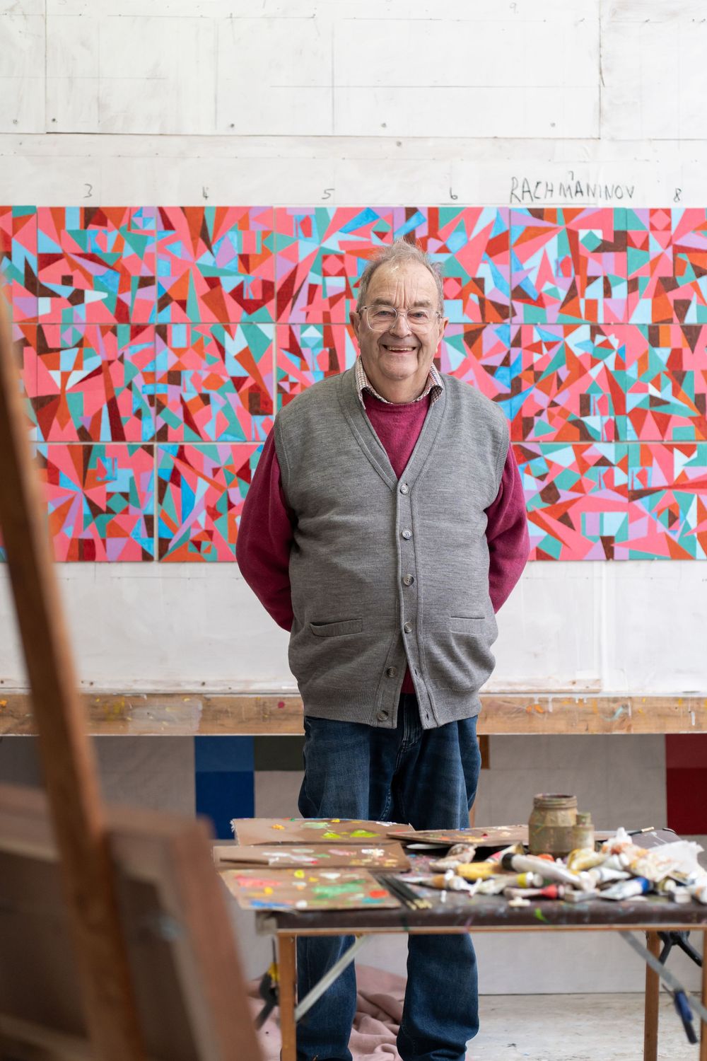 Smiling artist stands in front of colorful abstract painting in art studio with paint palette and brushes in foreground.